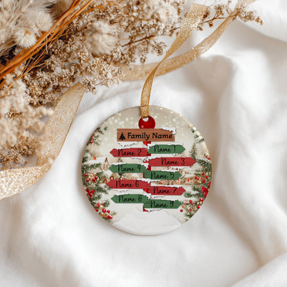 A round Christmas ornament with a personalized sign design, hanging from a gold ribbon on white fabric with dried flowers in the background.