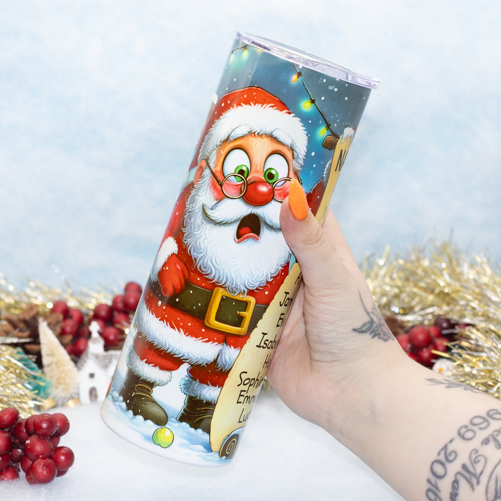 A hand holding a stainless steel tumbler with a colourful printed illustration of Santa Claus on a snowy night scene. The background is decorated with gold tinsel, red berries, and a light blue textured backdrop.