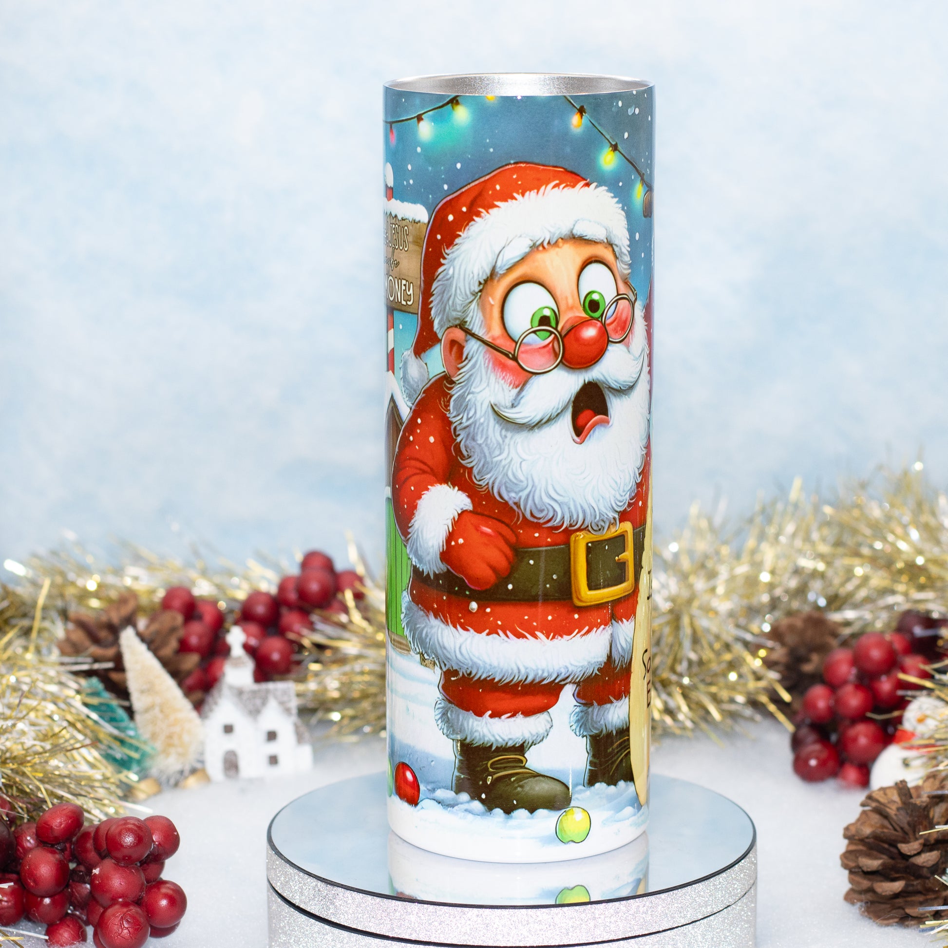 A cylindrical Christmas-themed tumbler featuring a cartoon Santa Claus standing in the snow with a sack on his shoulder, displayed on a silver pedestal with holiday decorations in the background.