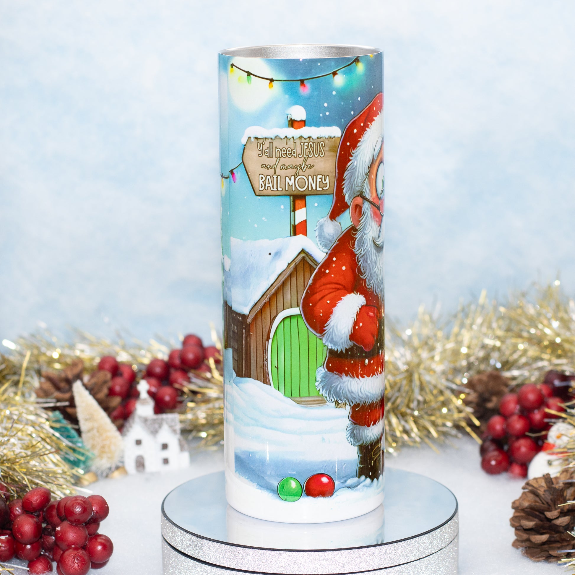 A cylindrical Christmas-themed tumbler displayed on a silver pedestal. The tumbler features a festive winter illustration with Santa Claus, a snow-covered cabin, a green door, colourful lights