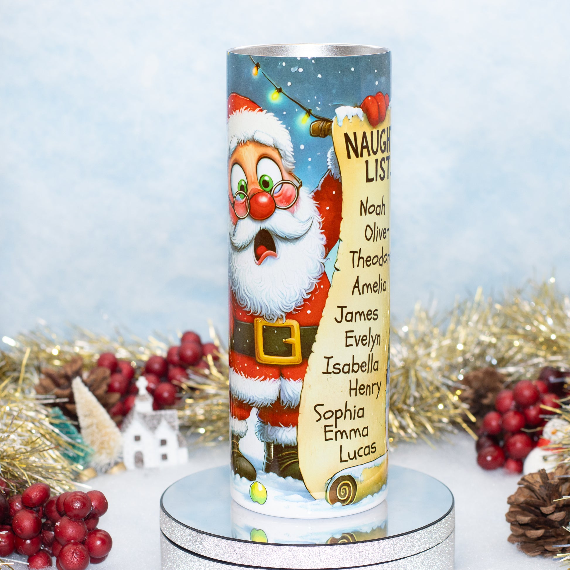 A Christmas cylindrical Tumbler featuring Santa Claus and a scroll with names under the heading 'NAUGHTY LIST'. The candle is displayed on a silver pedestal with festive holiday decorations in the background.
