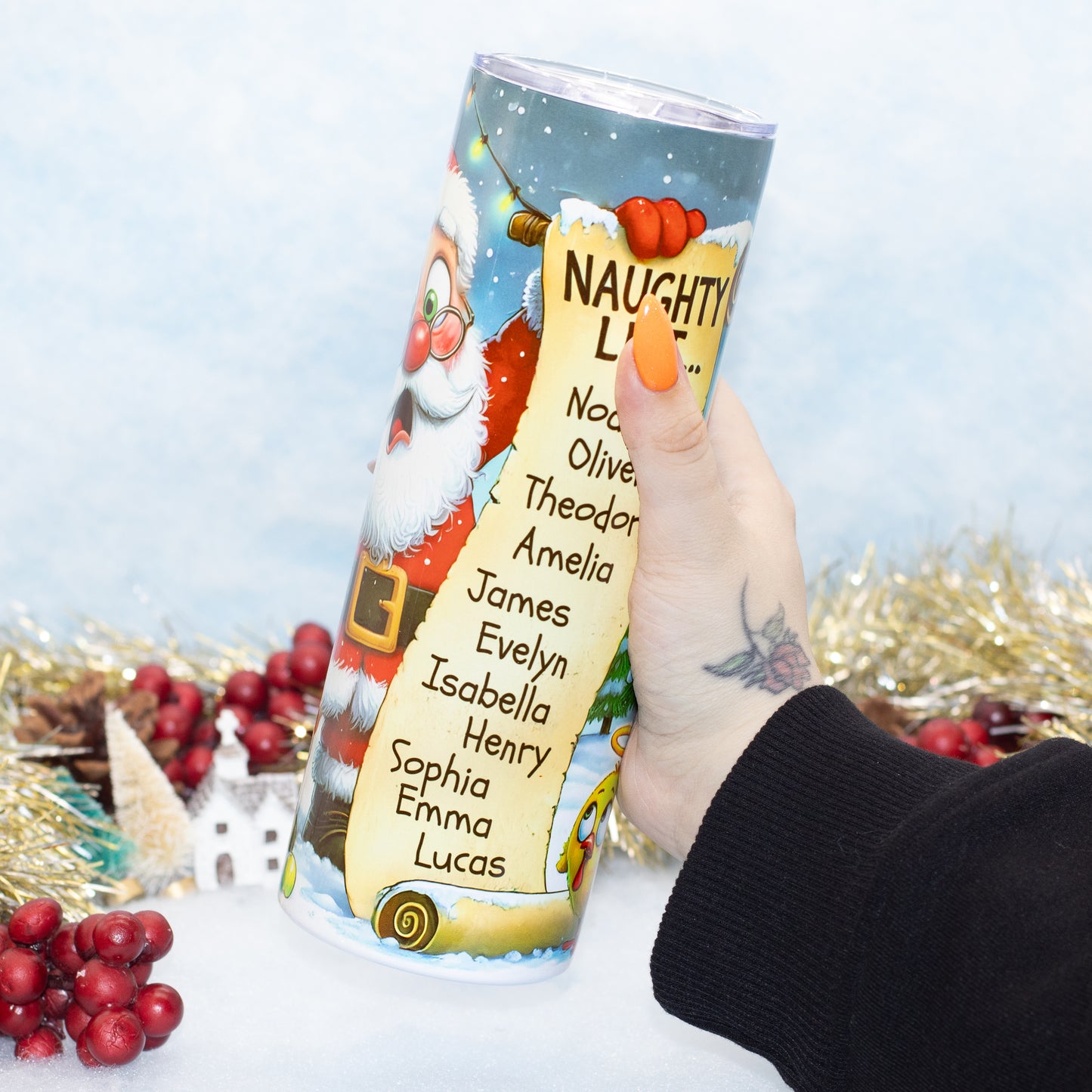 A hand holds a Christmas tumbler . The cylindrical tumbler features festive illustrations of Santa Claus and a golden scroll listing names, set against a snowy winter scene with gold tinsel and red berries in the background.
