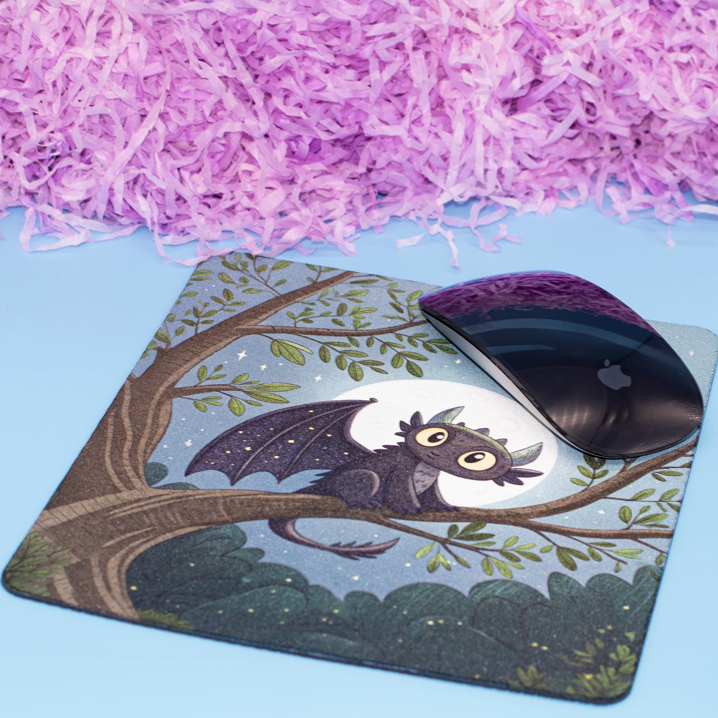 A square mouse pad with a printed illustration of a small bat perched on a tree branch at night, shown on a light blue surface with pink shredded paper in the background. A black computer mouse rests on the pad.