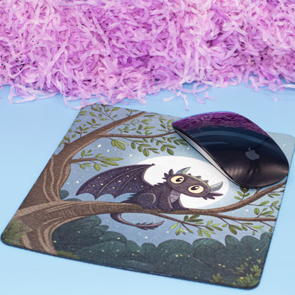 A square mouse pad with a printed illustration of a small bat perched on a tree branch at night, shown on a light blue surface with pink shredded paper in the background. A black computer mouse rests on the pad.