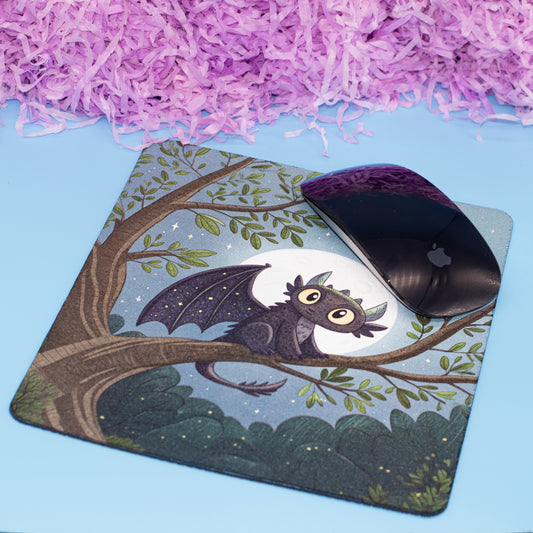 A rectangular desk mat with a printed illustration of a cartoon dragon perched on a tree branch under a full moon, shown with a black computer mouse on top. The mat is placed on a light blue surface with purple shredded paper in the background.