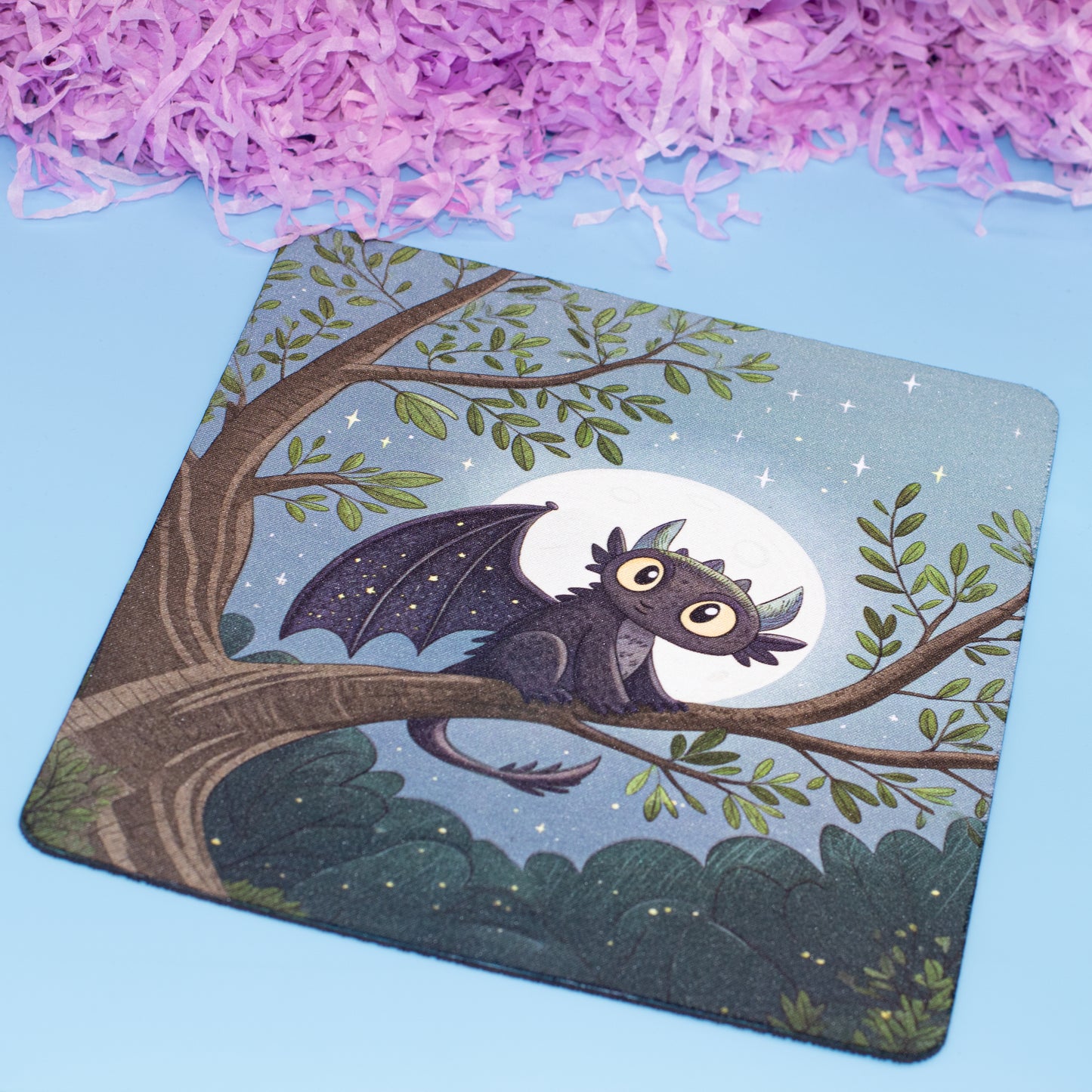 A square mouse pad with rounded corners featuring an illustration of a cute dark gray dragon perched on a tree branch under a full moon, set against a light blue background with decorative purple shredded paper at the top.