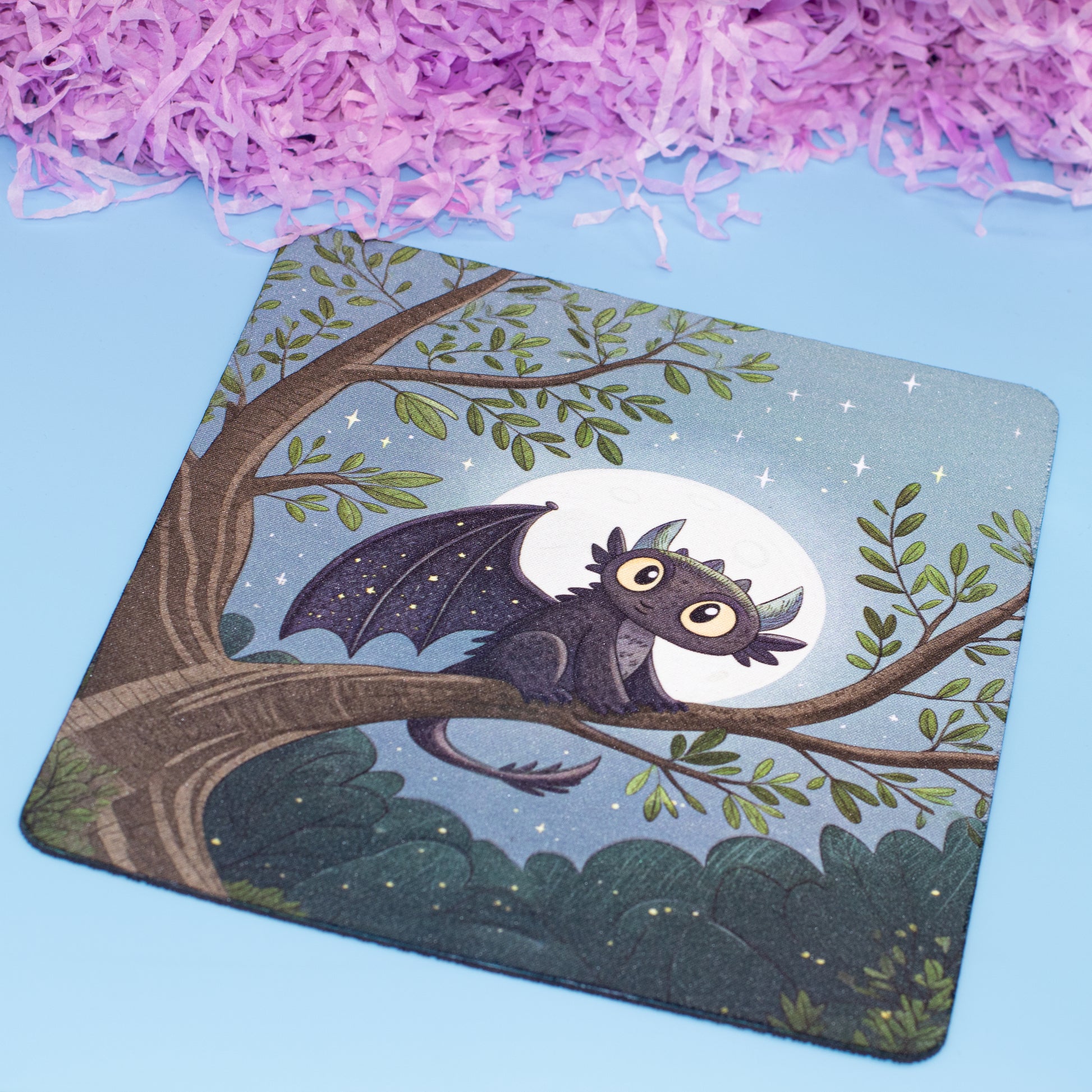 A square mouse pad with rounded corners featuring an illustration of a cute dark gray dragon perched on a tree branch under a full moon, set against a light blue background with decorative purple shredded paper at the top.