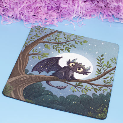 A square mouse pad with rounded corners featuring an illustration of a cute dark gray dragon perched on a tree branch under a full moon, set against a light blue background with decorative purple shredded paper at the top.