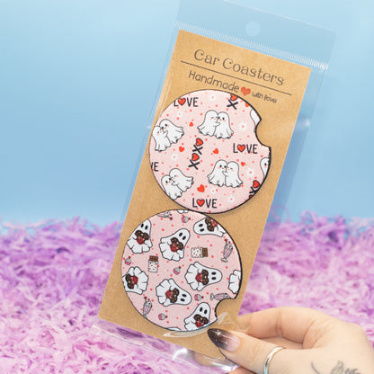 A hand holds a clear plastic package containing two round car coasters with a pink ghost and heart pattern on a kraft paper backing card. The background is a soft blue gradient with purple shredded paper filler.