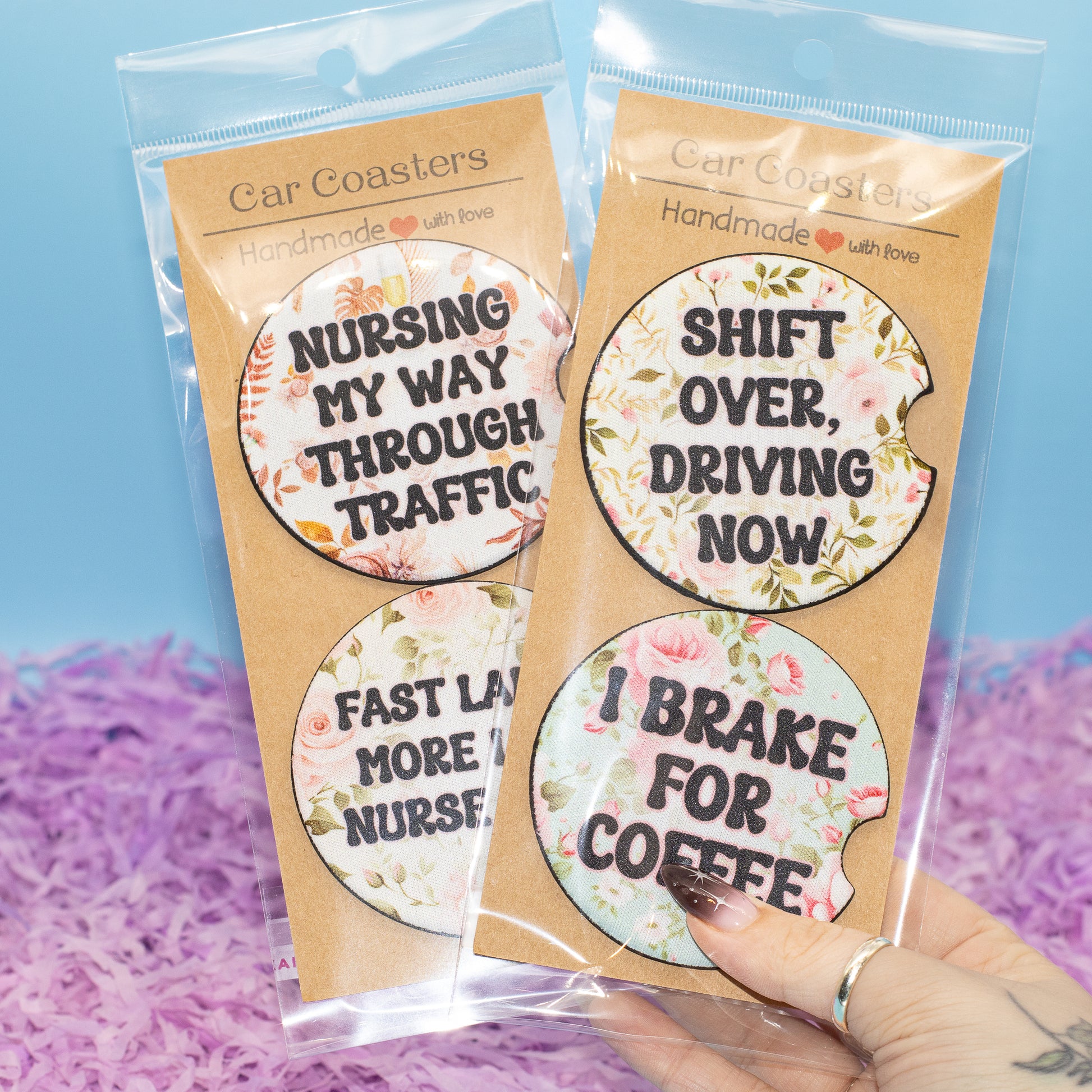 Two packaged car coasters with floral designs and humorous text, held by a hand against a blue and purple background.