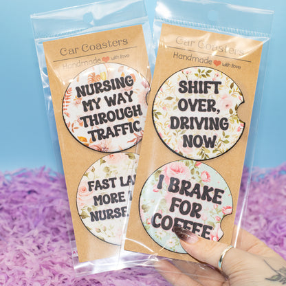Two packaged car coasters with floral designs and humorous text, held by a hand against a blue and purple background.