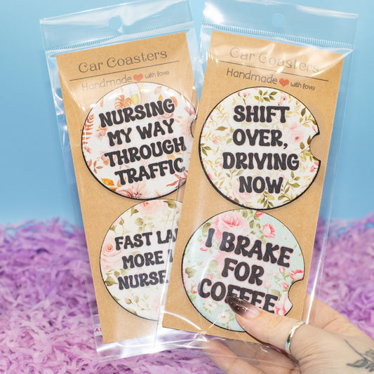 Two packaged car coasters with floral designs and humorous text, held by a hand against a blue and purple background.