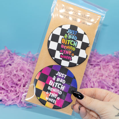 A hand  holds a clear plastic bag containing two round car coasters on a kraft card. The coasters have a black-and-white checkerboard design with the phrase 'JUST A BAD BITCH BEHIND THE WHEEL' in colourful lettering.