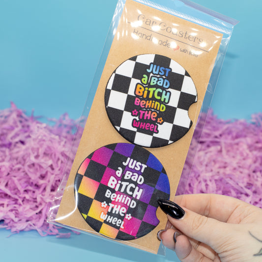 A hand  holds a clear plastic bag containing two round car coasters on a kraft card. The coasters have a black-and-white checkerboard design with the phrase 'JUST A BAD BITCH BEHIND THE WHEEL' in colourful lettering.