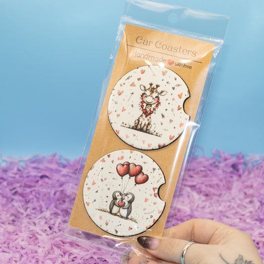 A hand holds a clear plastic package containing two round car coasters. The coasters have a white background with tiny red hearts and illustrated animals: a giraffe with a floral wreath and a couple holding red heart-shaped balloons.