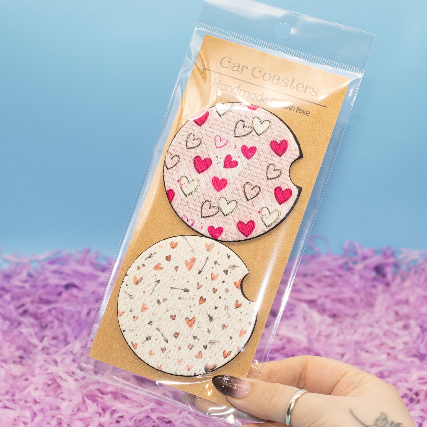 A hand holds a clear plastic package containing two round car coasters with heart patterns. The top coaster is light pink with red and black hearts, and the bottom coaster is white with peach and black hearts.
