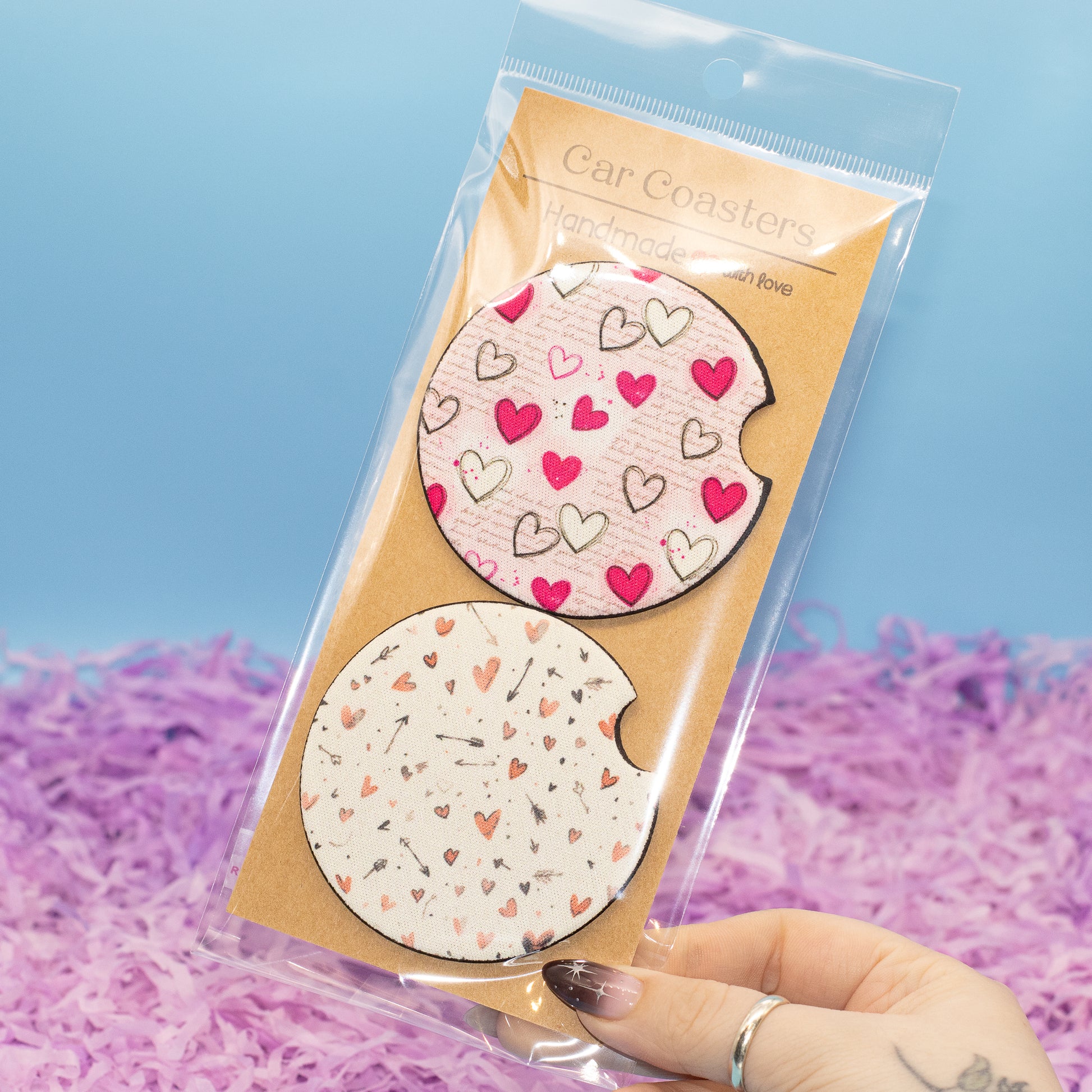A hand holds a clear plastic package containing two round car coasters with heart patterns. The top coaster is light pink with red and black hearts, and the bottom coaster is white with peach and black hearts.