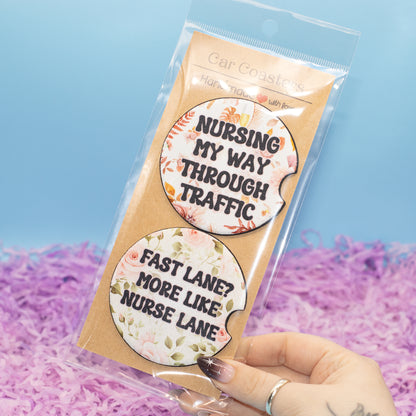 A hand holds a clear plastic bag containing two oval car coasters with floral designs and humorous text, set against a light blue background with purple shredded paper filler.