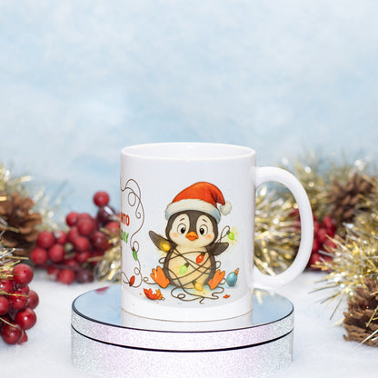 A white ceramic mug featuring a cute cartoon penguin wearing a Santa hat and wrapped in Christmas lights, displayed on a silver round pedestal with festive holiday decorations in the background.
