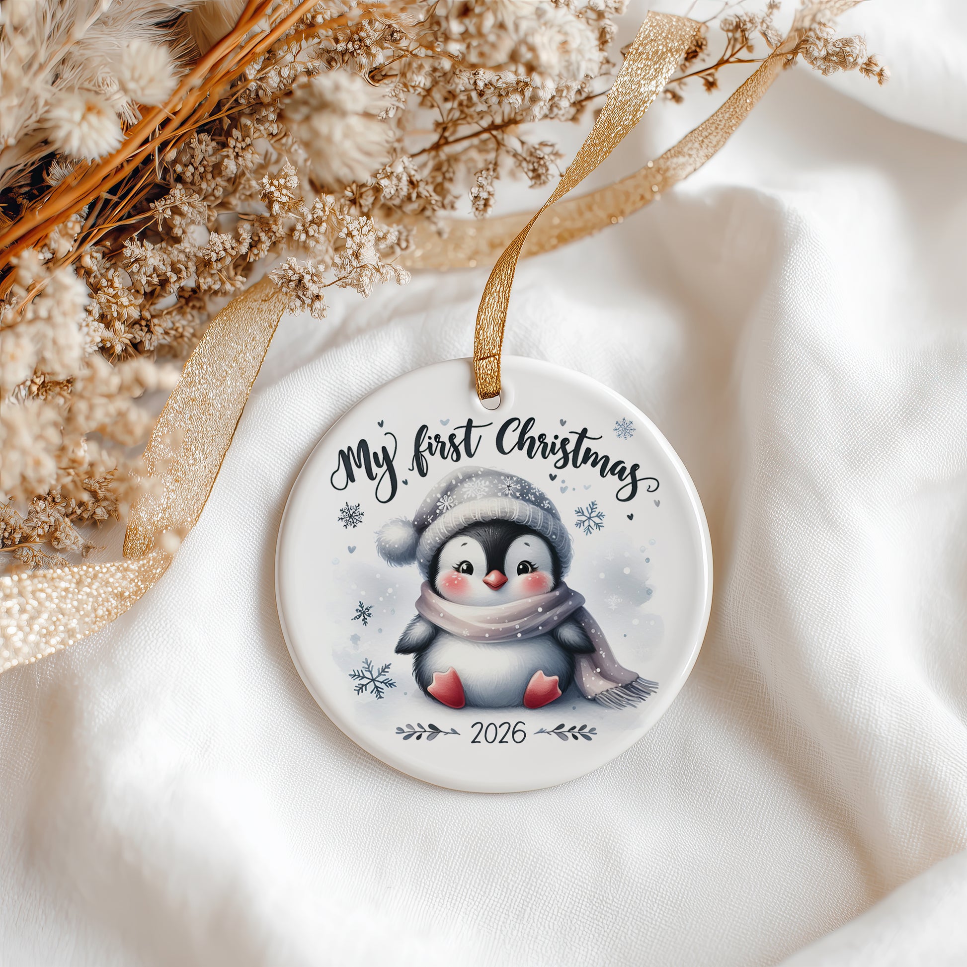 A round white ceramic Christmas ornament featuring a cute penguin illustration and the text 'My first Christmas 2026'. The ornament hangs from a gold ribbon on a soft white fabric background with dried flowers and a gold ribbon.