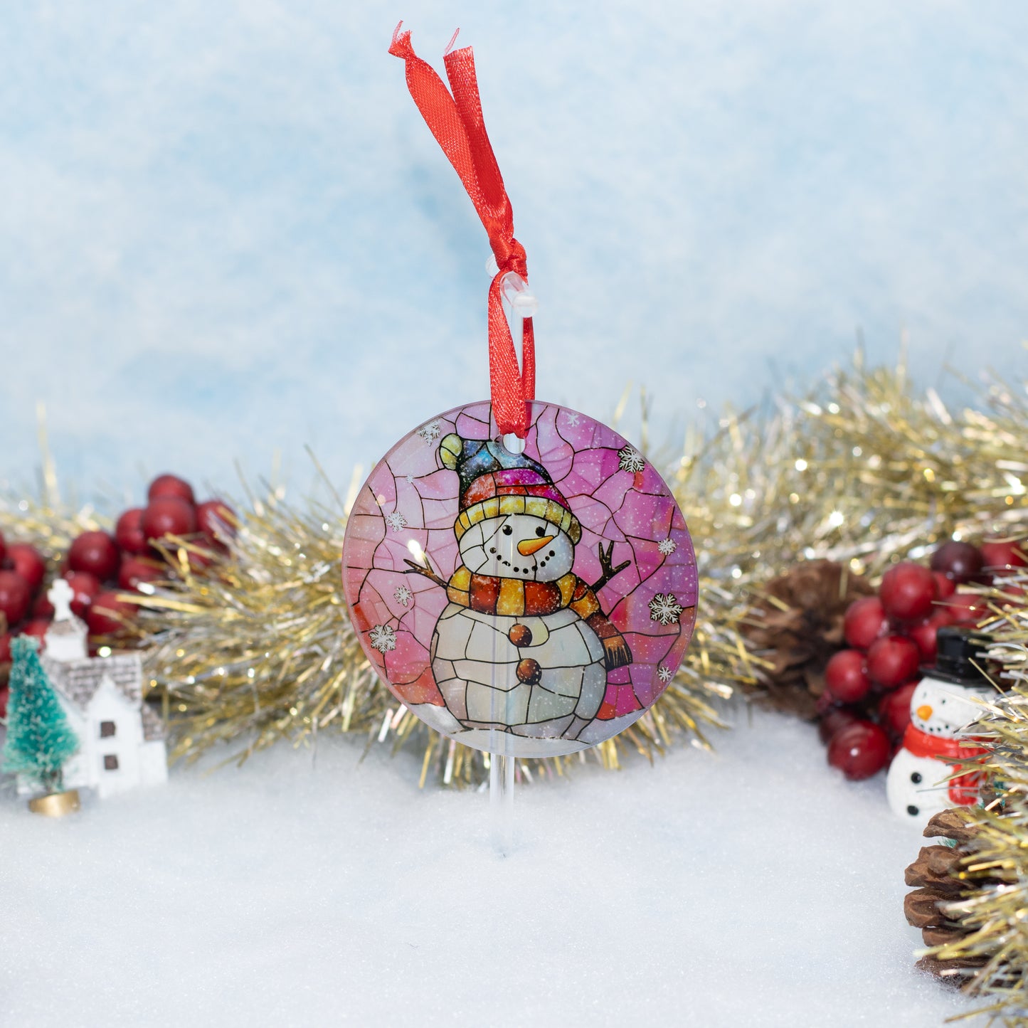 A round Christmas ornament featuring a painted snowman under a pink and purple cracked-glass style sky, hanging from a red ribbon. The ornament is styled on a snowy surface with gold tinsel, red berries, pinecones, and miniature holiday decorations in the background.