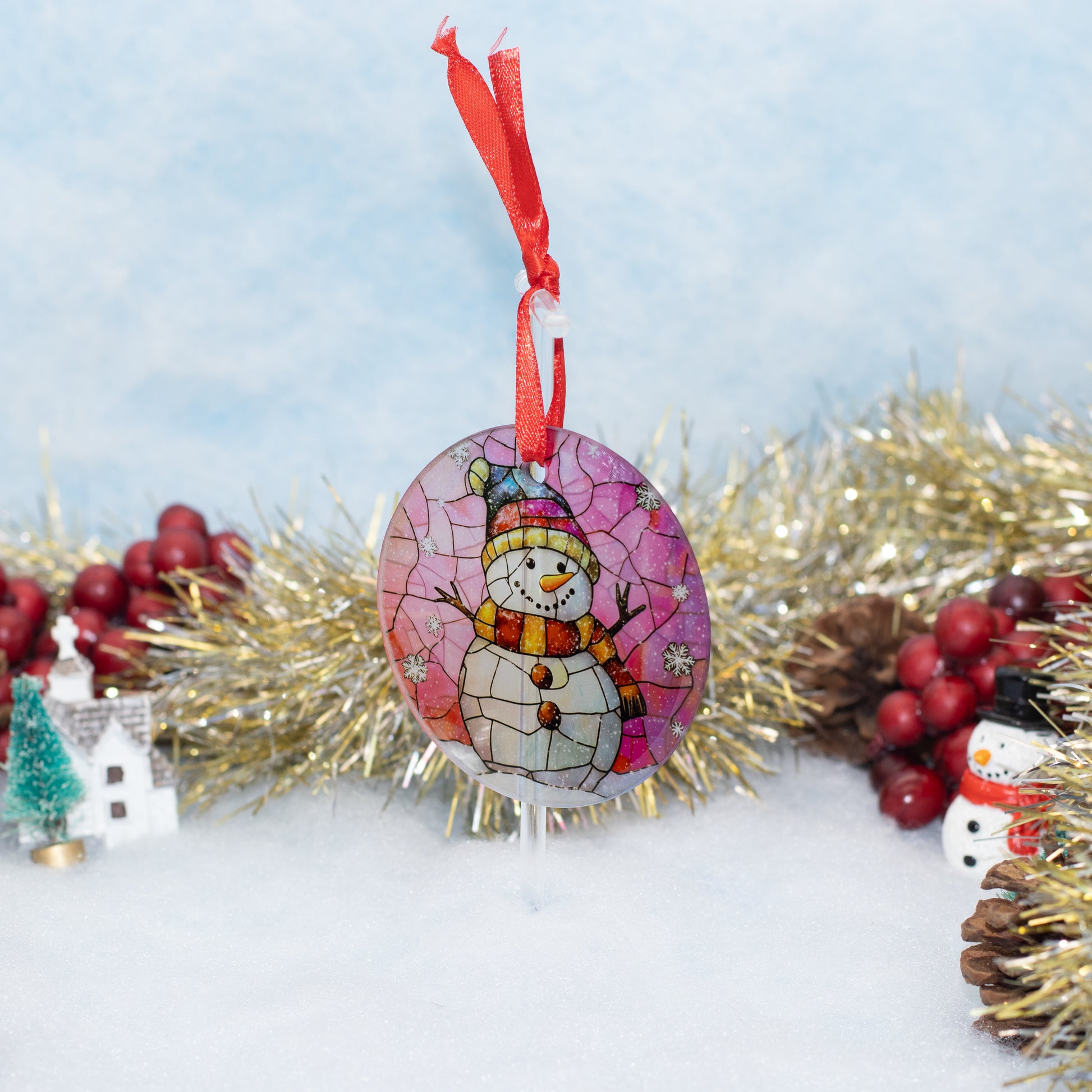A round Christmas ornament featuring a stained-glass style snowman wearing a blue hat and orange scarf, hanging from a red ribbon. The ornament is displayed on a white surface with festive decorations including gold tinsel, red berries, pinecones, and miniature houses against a soft light blue background.