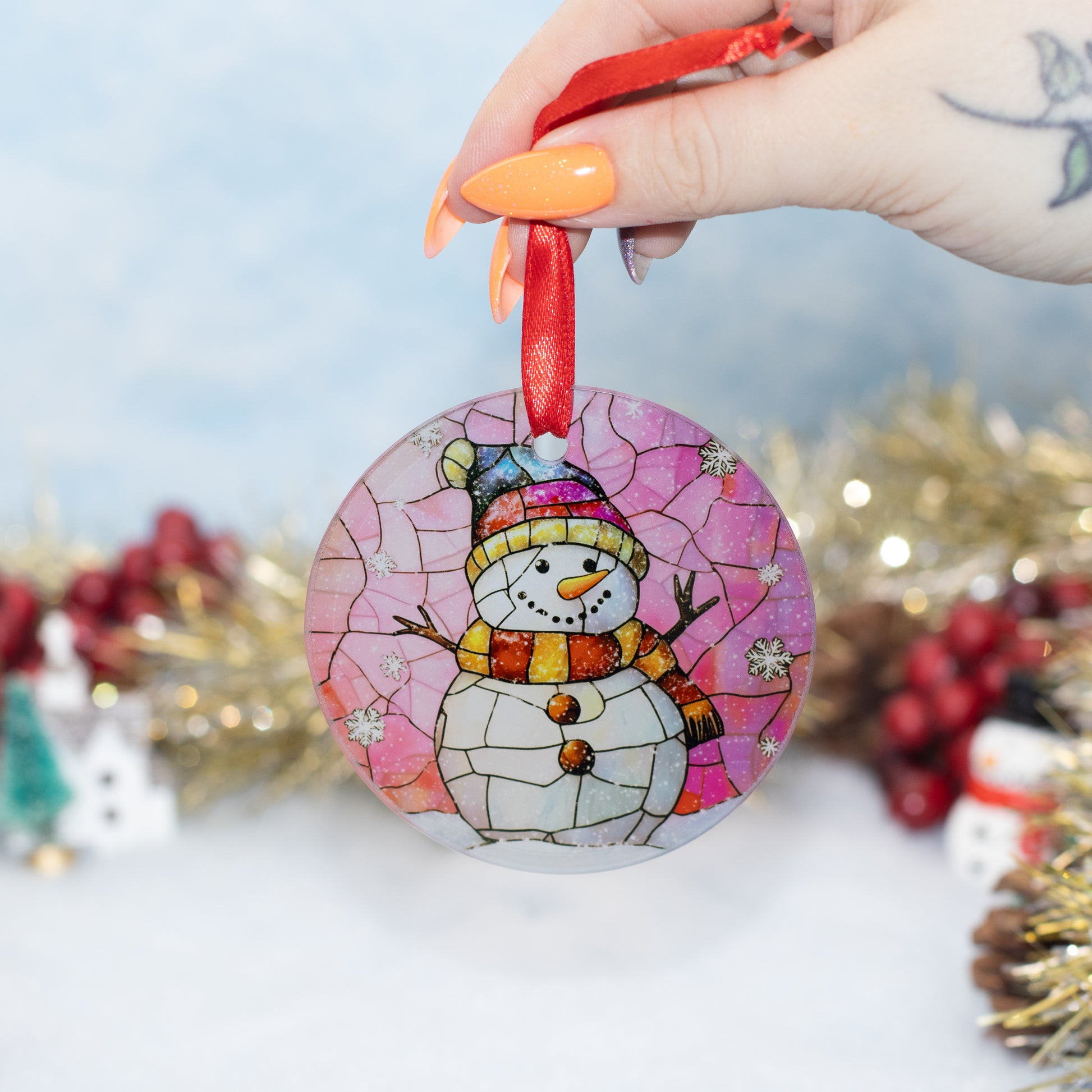 A hand holds a round stained-glass style Christmas ornament featuring a snowman wearing a pink and multicolour hat against a pink background. The ornament has a red hanging ribbon and is photographed in a festive setting with blurred gold tinsel, red berries, and small holiday decorations in the background.