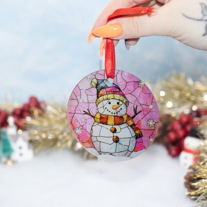 A hand holds a round stained-glass-style Christmas ornament featuring a snowman on a pink background with a red hanging ribbon. The scene is softly blurred with holiday decorations in the background.