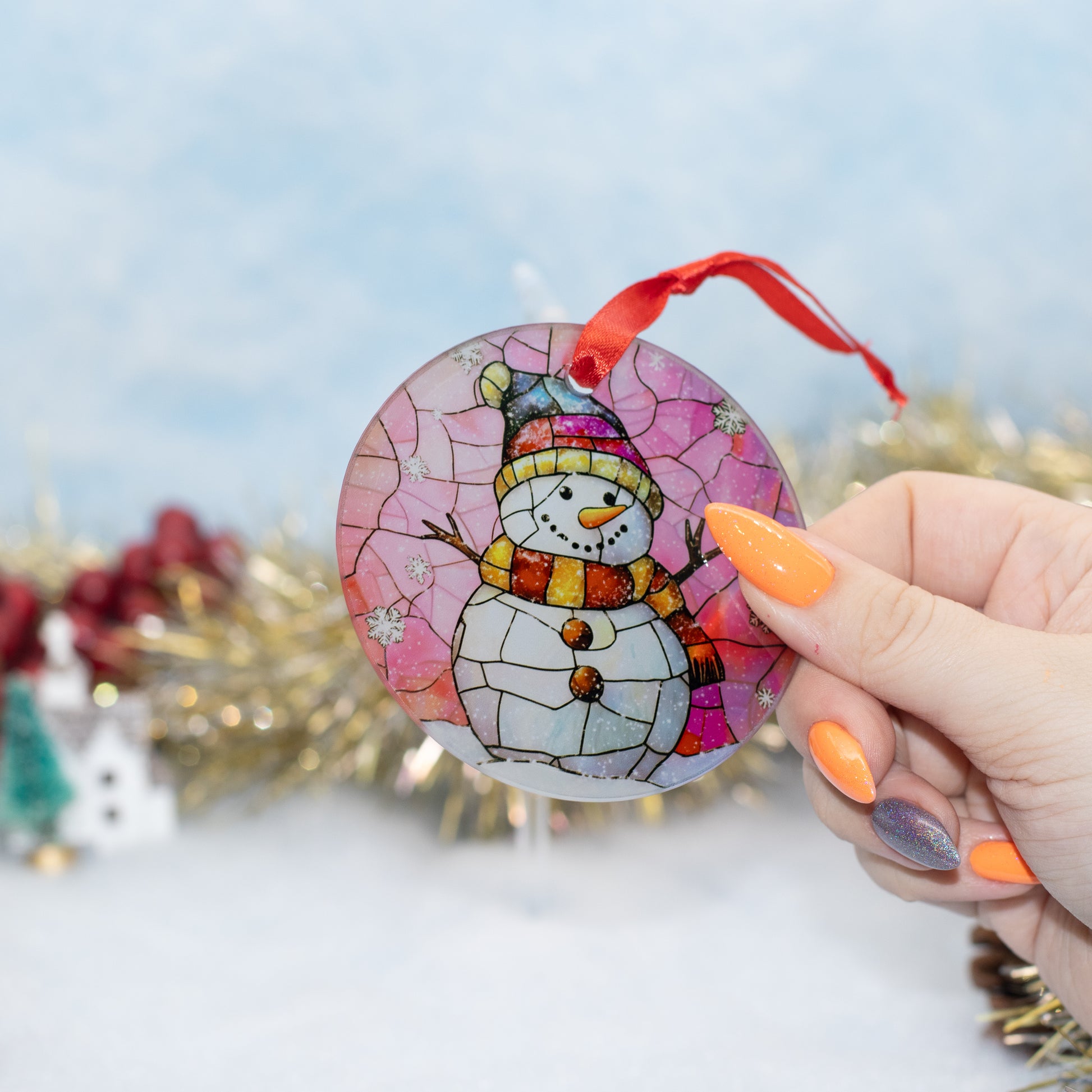 A hand holds a round stained-glass-style snowman ornament with a red ribbon hanger against a blurred festive background.