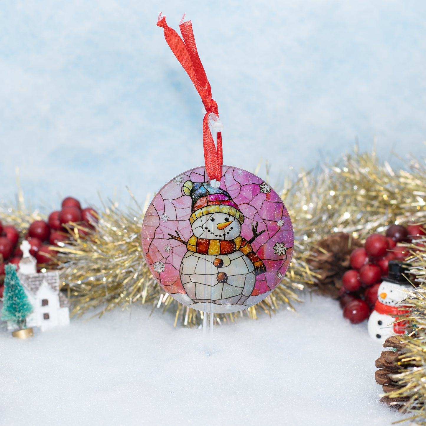 A round stained-glass style snowman Christmas ornament with a red ribbon hanger, displayed on a snowy surface with gold tinsel, red berries, and small holiday decorations in the background.