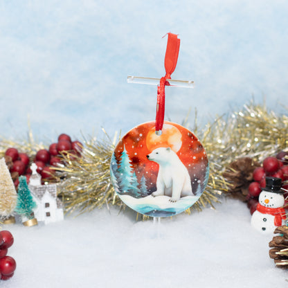 A round Christmas ornament featuring a painted polar bear in a snowy winter scene, hanging from a red ribbon on a clear stand. The ornament is displayed in a festive holiday setting with gold tinsel, red berries, a small house, and a snowman against a soft light blue background.