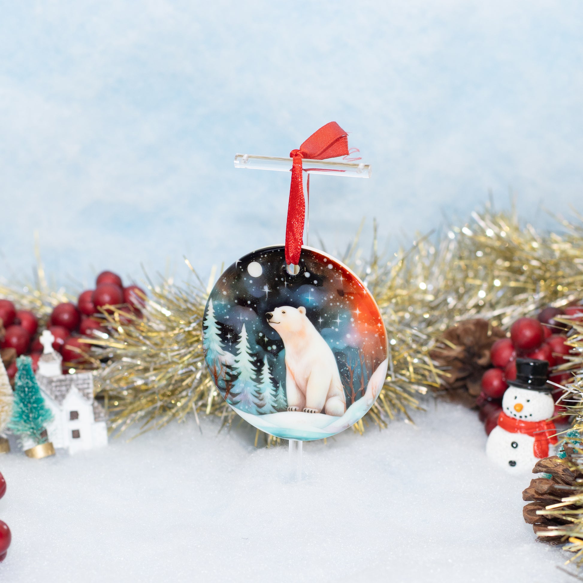 A round Christmas ornament featuring an illustration of a polar bear in a snowy night landscape, hanging from a clear stand with a red ribbon. The ornament is styled on a white surface with festive decorations including gold tinsel, red berries, a small house, and a snowman against a soft blue background.
