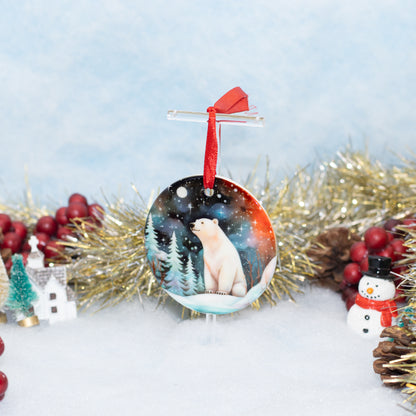 A round Christmas ornament featuring an illustration of a polar bear in a snowy night landscape, hanging from a clear stand with a red ribbon. The ornament is styled on a white surface with festive decorations including gold tinsel, red berries, a small house, and a snowman against a soft blue background.