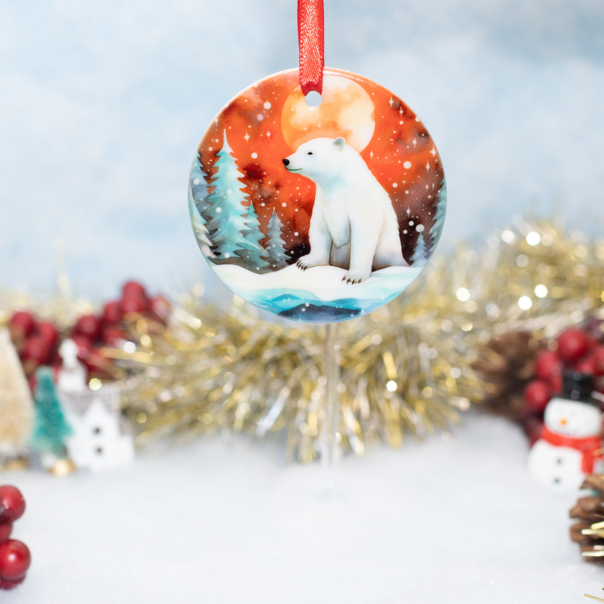 A round Christmas ornament featuring a painted polar bear in a snowy winter scene, hanging by a red ribbon against a softly blurred holiday background with tinsel and red berries.