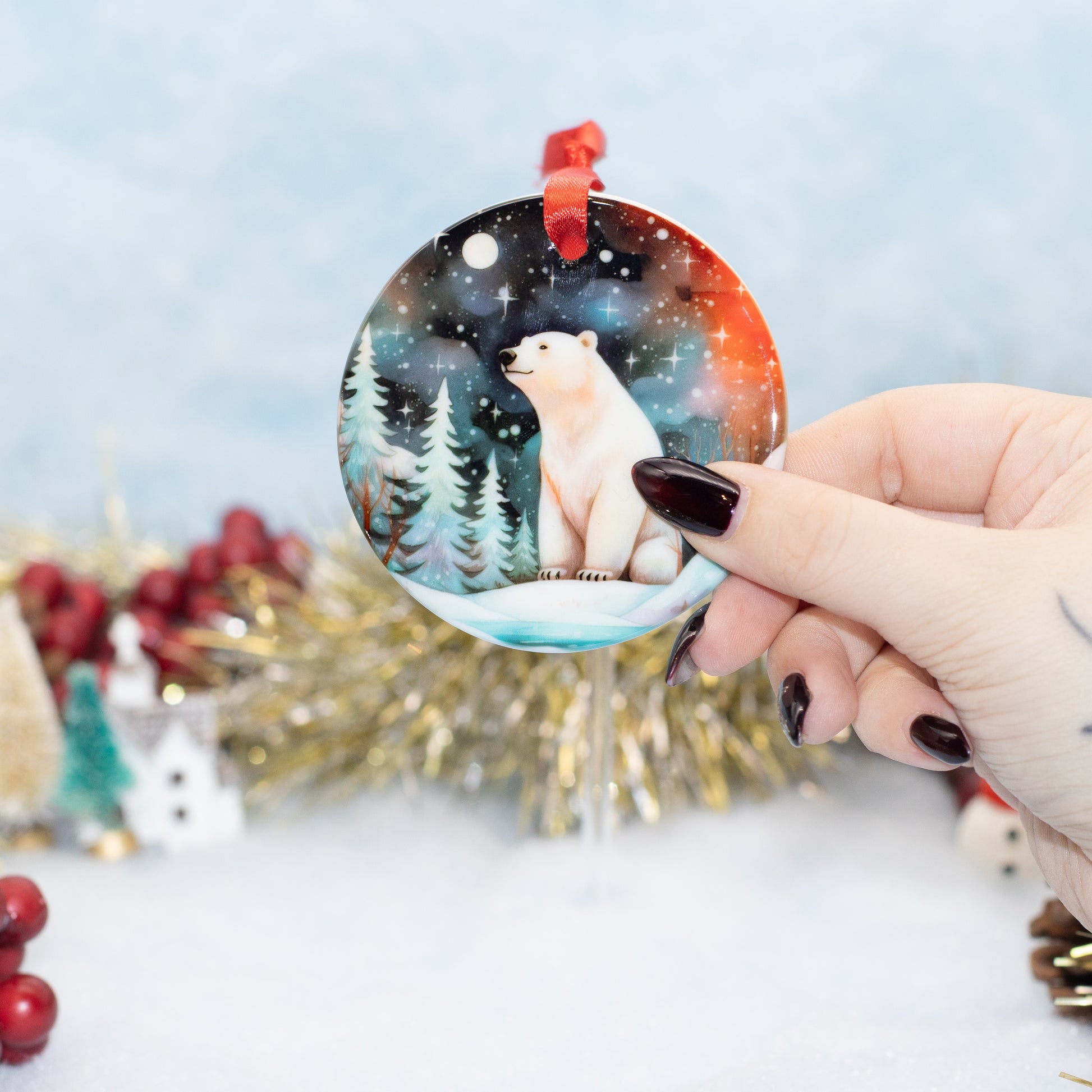 A hand holds a round Christmas ornament featuring a painted polar bear in a snowy night landscape. The ornament has a red hanging loop and is shown against a softly blurred festive background with tinsel and holiday decorations.