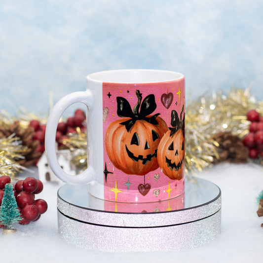 A white ceramic mug with a pink Halloween-themed illustration of two jack-o'-lantern pumpkins, displayed on a round silver pedestal with festive holiday decorations in the background.