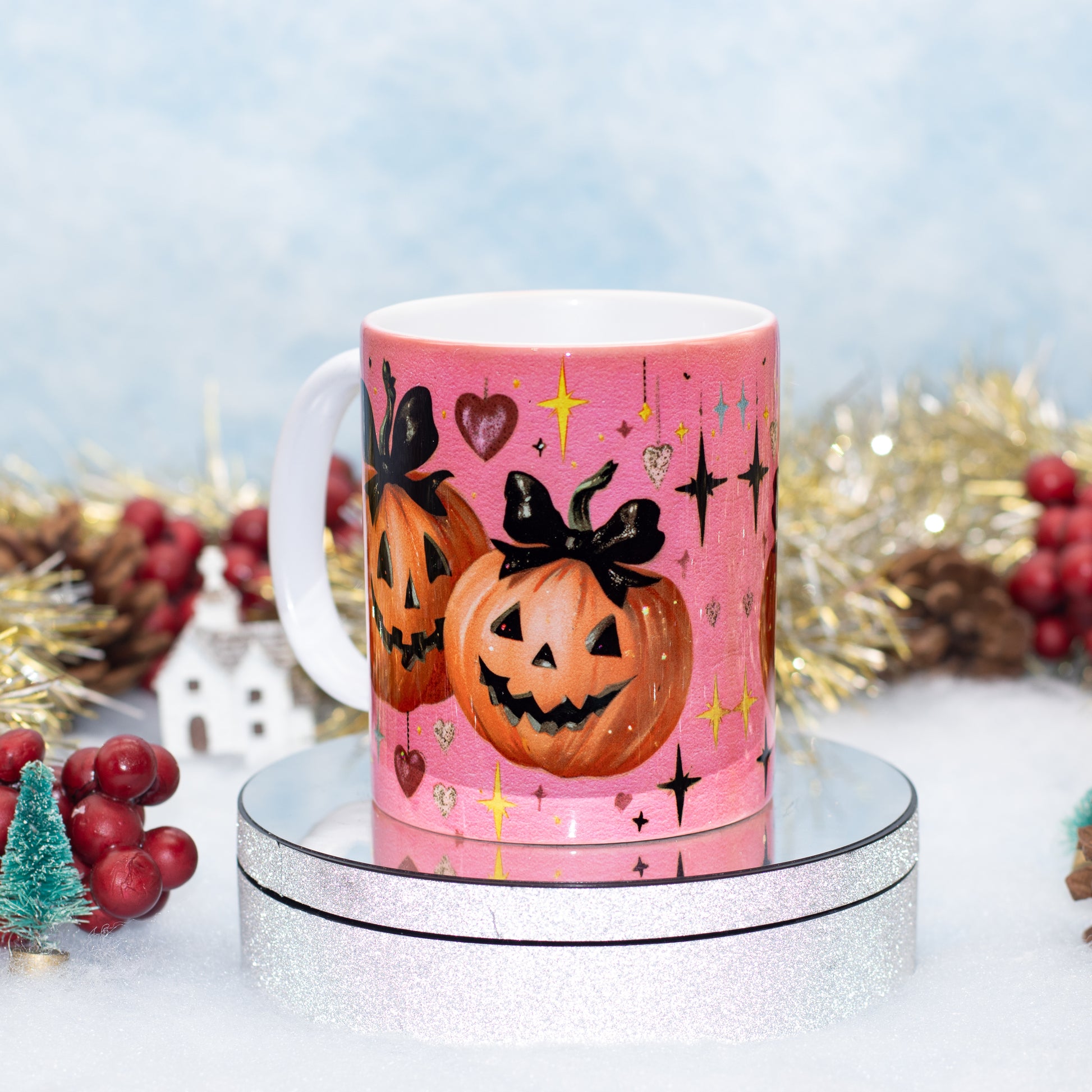 A pink ceramic mug with a Halloween design, featuring two jack-o'-lanterns with black bows, hearts, stars, and sparkles, displayed on a silver glitter pedestal with festive holiday decorations in the background.