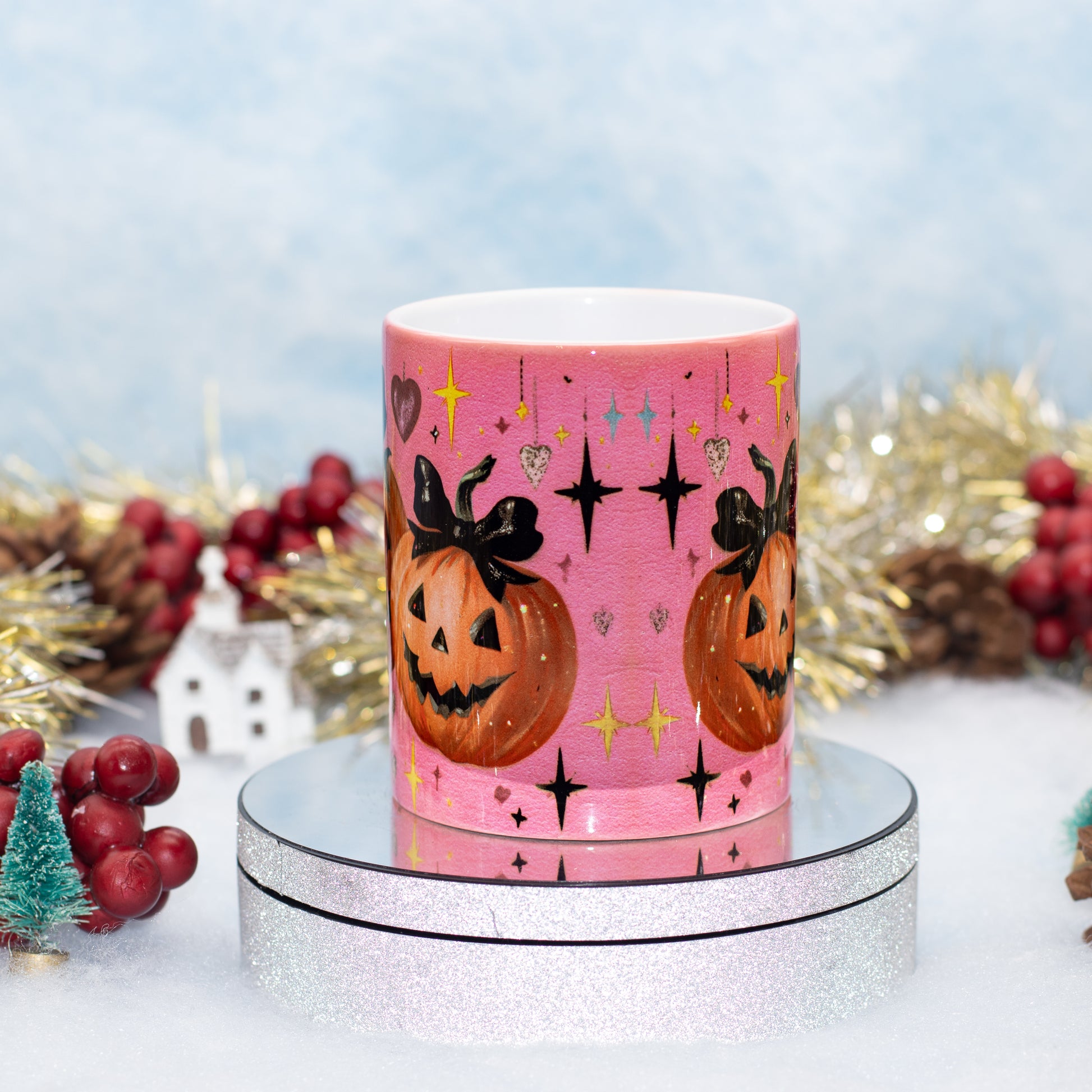 A ceramic mug with a Halloween-themed illustrations of two jack-o'-lanterns, black bows, stars, and sparkles, displayed on a round silver pedestal with festive holiday decorations in the background.