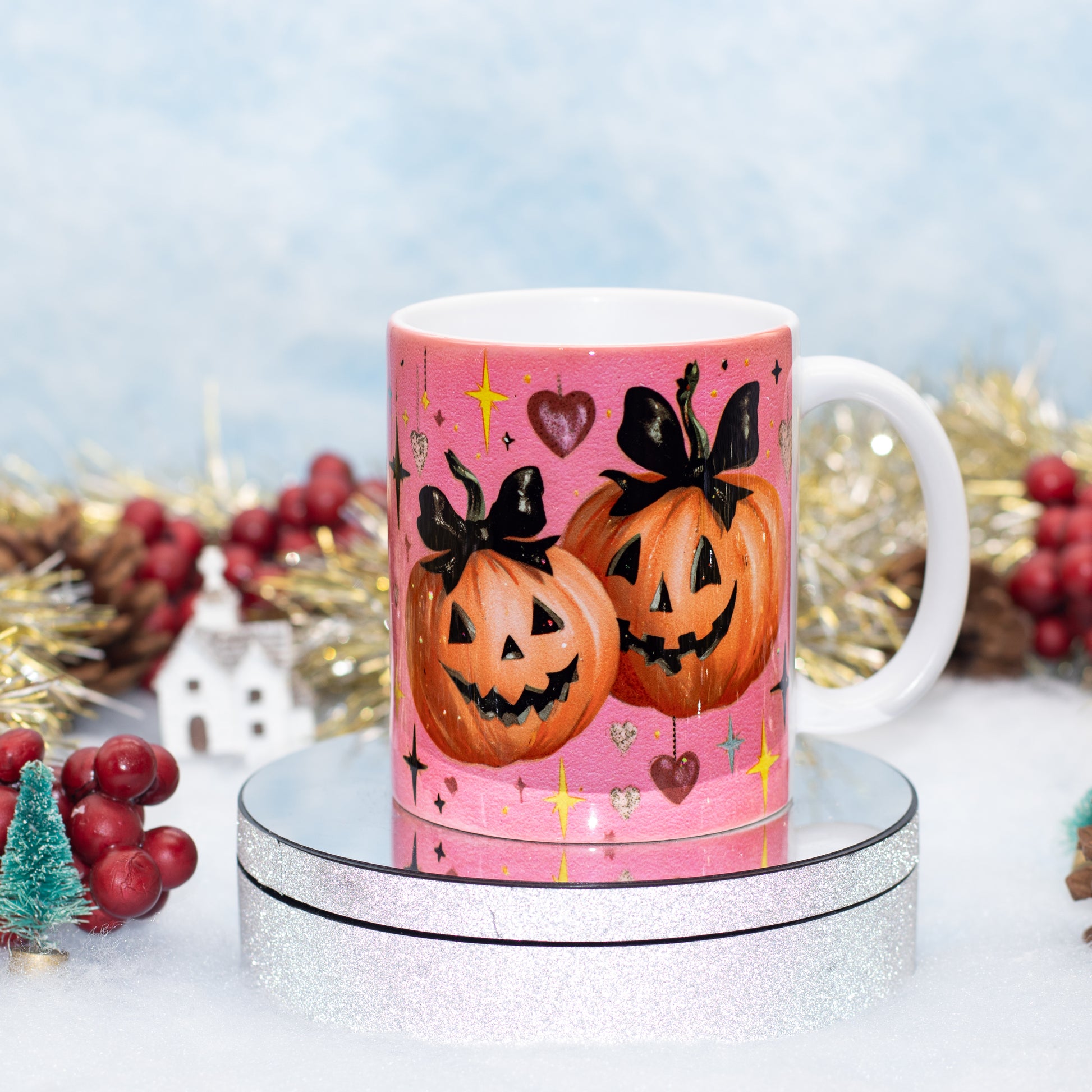 A white ceramic mug with a pink Halloween design featuring two jack-o'-lantern pumpkins with black bows, displayed on a silver glitter stand with festive holiday decorations in the background.