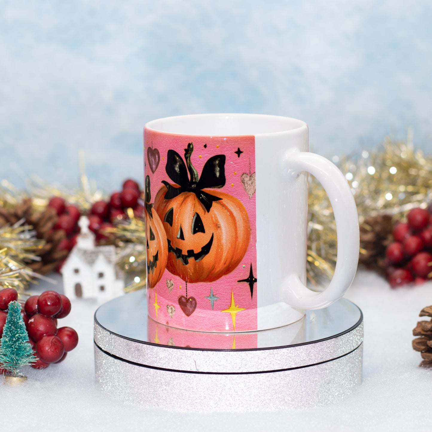 A white ceramic mug with a pink Halloween-themed print featuring jack-o'-lantern pumpkins, hearts, and stars, displayed on a round silver pedestal with festive holiday decorations in the background.