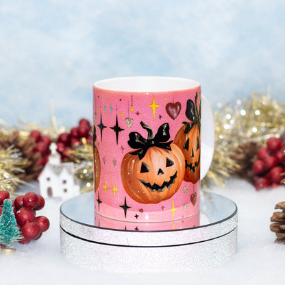 A pink ceramic mug decorated with jack-o'-lantern pumpkins, black bows, and gold stars, displayed on a silver glitter stand with festive holiday decorations in the background.