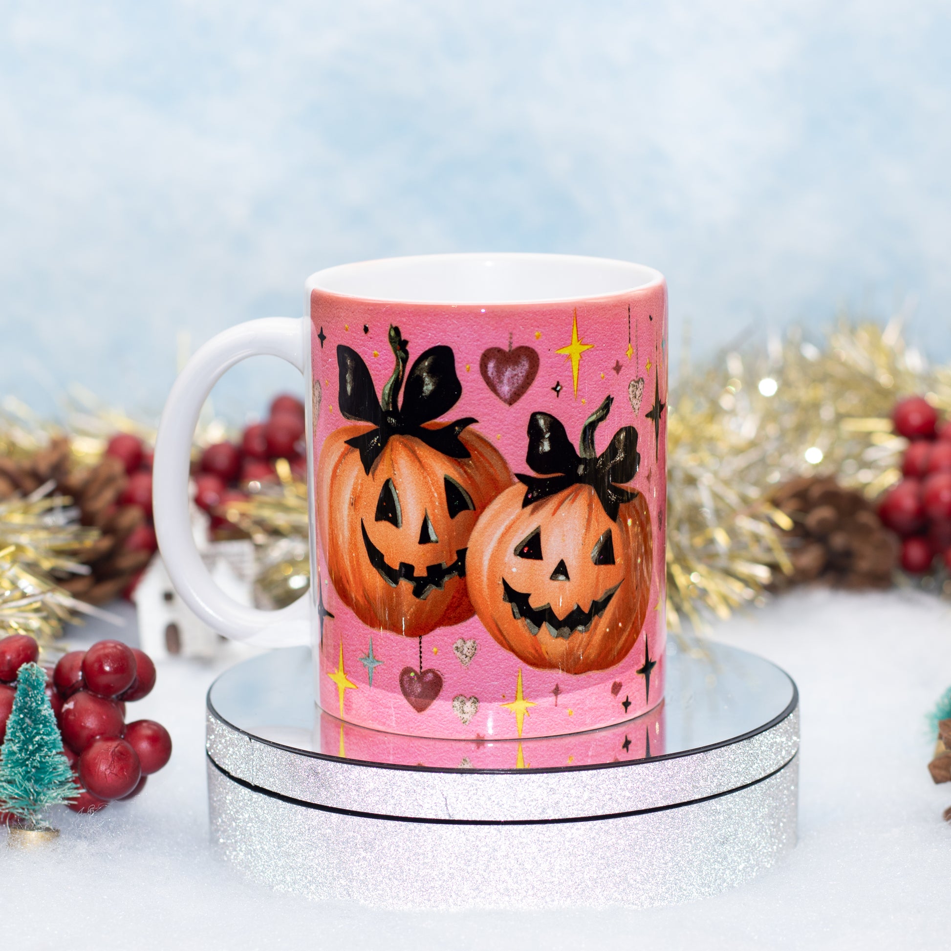 A ceramic mug with a Halloween design featuring two jack-o'-lanterns wearing black bows, displayed on a round silver pedestal with festive holiday decorations in the background.