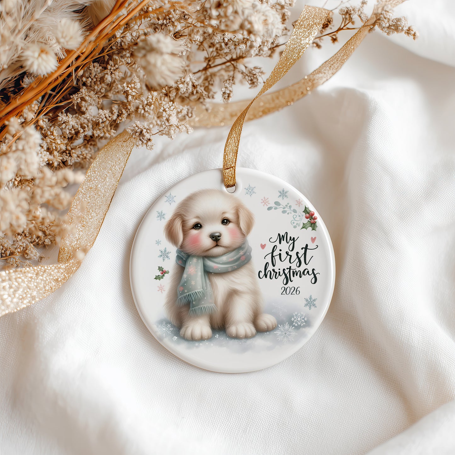 A round ceramic Christmas ornament featuring an illustration of a puppy wearing a blue scarf, with the text 'My first Christmas 2026' and small snowflakes. The ornament hangs from a gold ribbon and is styled on white fabric with dried flowers and a gold ribbon.