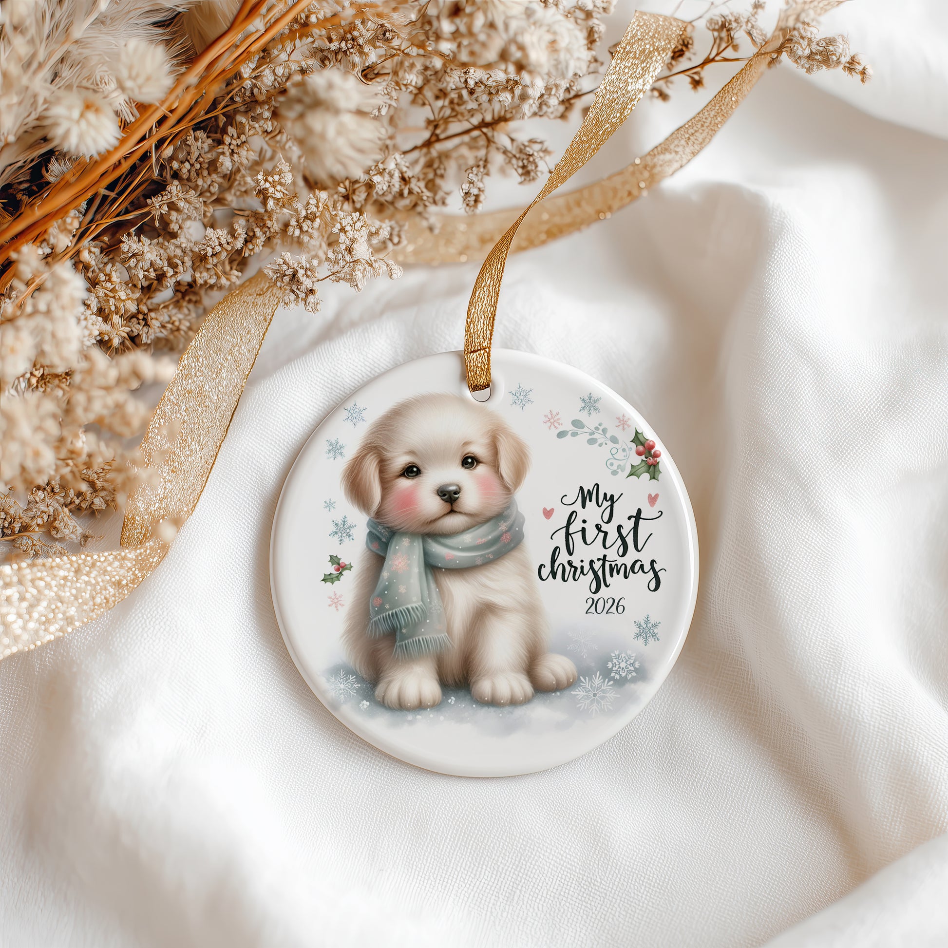 A round ceramic Christmas ornament featuring an illustration of a puppy wearing a blue scarf, with the text 'My first Christmas 2026' and small snowflakes. The ornament hangs from a gold ribbon and is styled on white fabric with dried flowers and a gold ribbon.