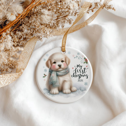 A round ceramic Christmas ornament featuring an illustration of a puppy wearing a blue scarf, with the text 'My first Christmas 2026' and small snowflakes. The ornament hangs from a gold ribbon and is styled on white fabric with dried flowers and a gold ribbon.