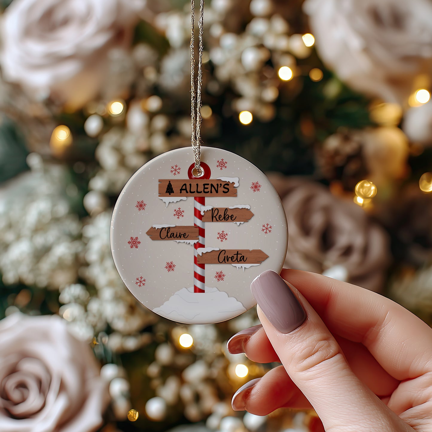 A round personalized Christmas ornament held by a hand, featuring a printed signpost with names, set against a blurred festive background with lights and roses.