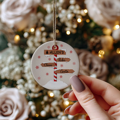 A round personalized Christmas ornament held by a hand, featuring a printed signpost with names, set against a blurred festive background with lights and roses.