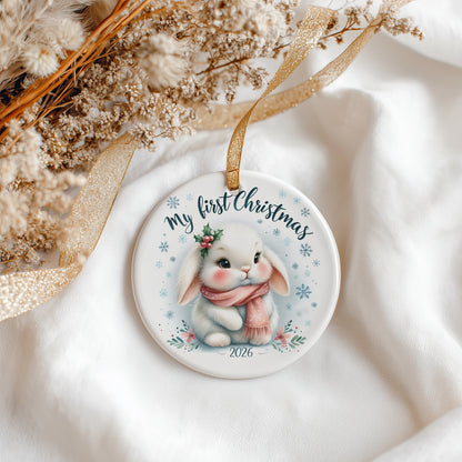 A round ceramic Christmas ornament featuring a cute white bunny wearing a pink scarf and holly, with the text 'my first Christmas' and '2026'. The ornament hangs from a gold ribbon and is styled on white fabric with dried flowers.