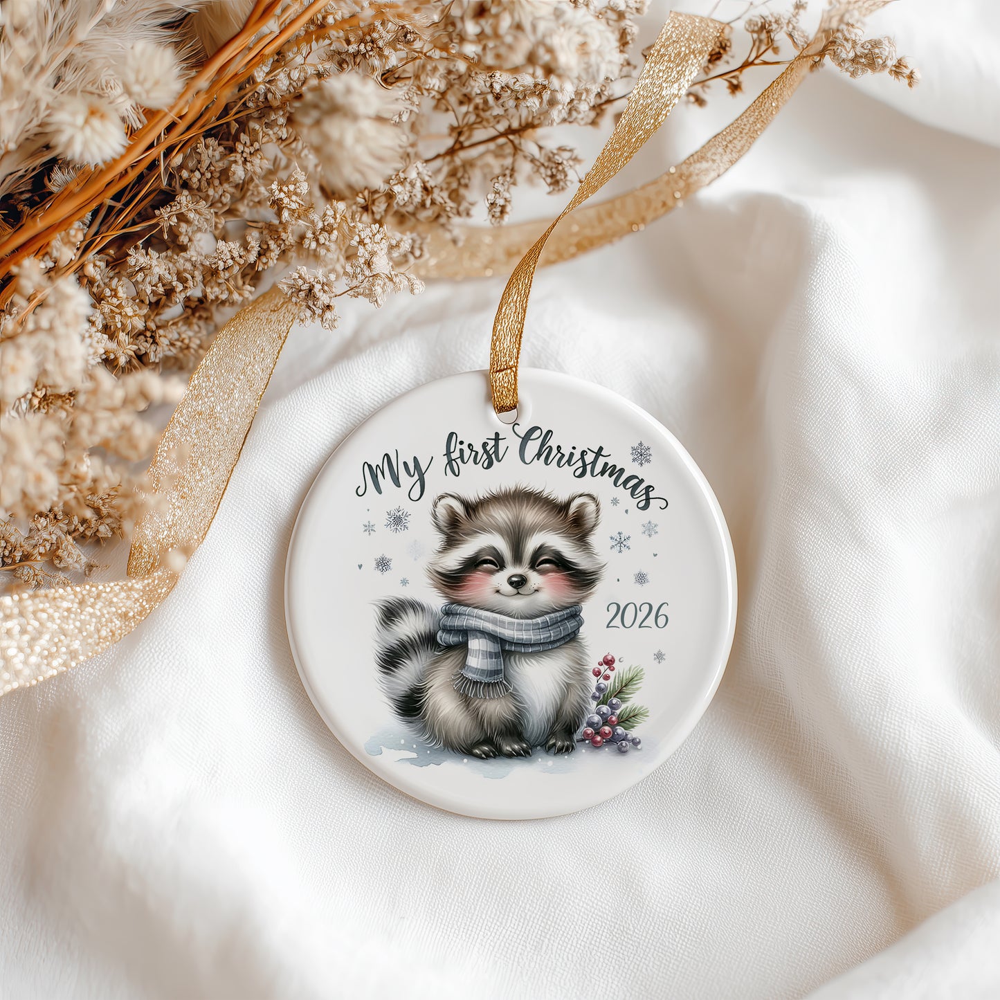 A round ceramic Christmas ornament featuring a cute illustrated raccoon wearing a grey scarf, with the text 'My first Christmas 2026' and small snowflake details. The ornament hangs from a gold ribbon on a soft white fabric background with dried flowers and a gold ribbon as styling props.