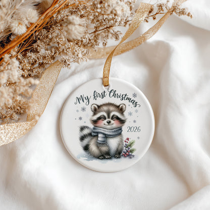 A round ceramic Christmas ornament featuring a cute illustrated raccoon wearing a grey scarf, with the text 'My first Christmas 2026' and small snowflake details. The ornament hangs from a gold ribbon on a soft white fabric background with dried flowers and a gold ribbon as styling props.